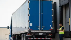 A blue semi-truck trailer without logos parked at a loading dock with a worker in a high-visibility vest standing nearby checking a handheld device.