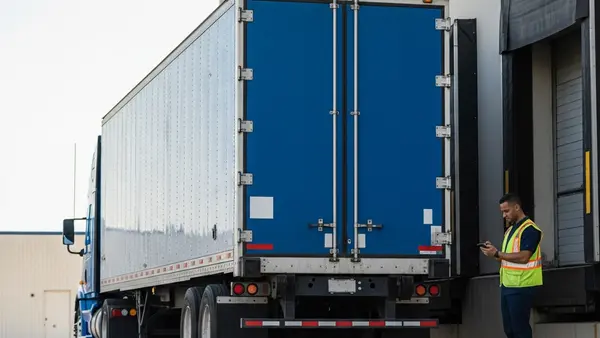 A blue semi-truck trailer without logos parked at a loading dock with a worker in a high-visibility vest standing nearby checking a handheld device.