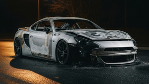 A charred silver sports car with significant front-end damage resting on a dark asphalt road at night near a street light.
