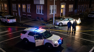 A patrol vehicle with flashing blue and red lights parked on a dark city street next to yellow caution tape at a crash site.