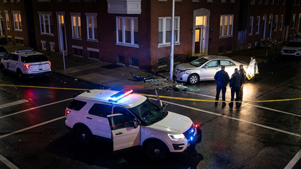 A patrol vehicle with flashing blue and red lights parked on a dark city street next to yellow caution tape at a crash site.