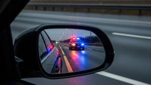 A police cruiser's flashing blue and red lights reflected in the rearview mirror of a stopped sedan on the side of a highway at dusk