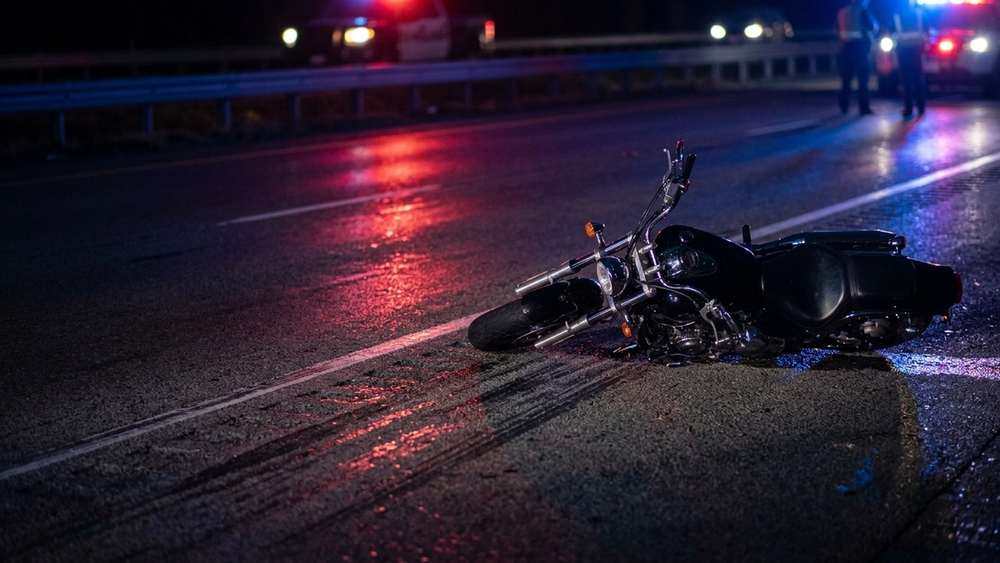 A dark-colored motorcycle lying on its side on a highway asphalt shoulder at night with blurred emergency lights reflecting on the pavement.