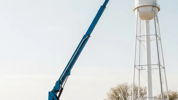 A large blue articulated boom lift extended high into the air toward a water tower on a construction site.