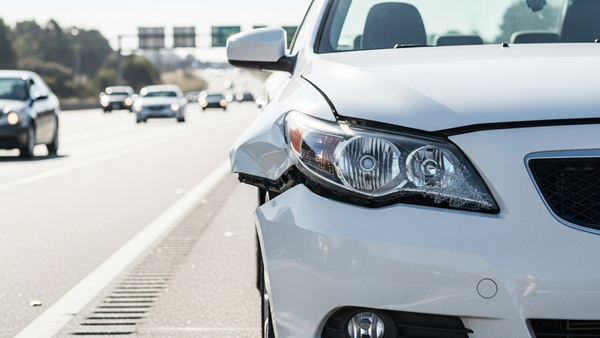 A side-view of a damaged white sedan on a California highway shoulder with traffic moving in the background, focusing on the crumpled fender and shattered headlight.