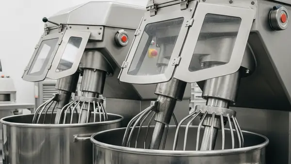 A large industrial dough mixing machine in a commercial bakery setting with safety guards and mechanical arms.