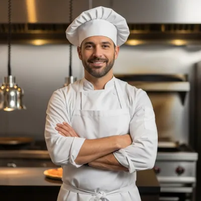 A pizza chef in a white apron stands in a restaurant kitchen with blurry stainless steel appliances in the background.