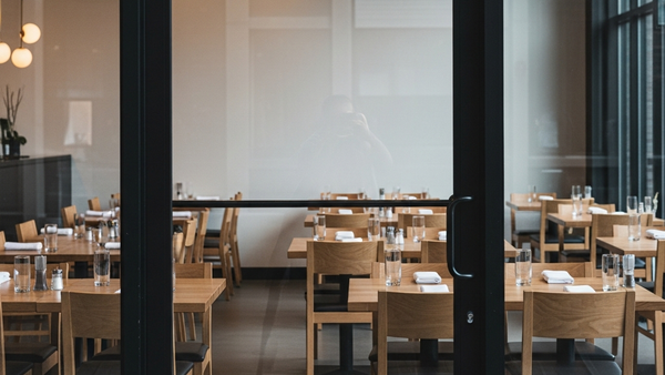 A modern restaurant dining room with empty wooden tables and chairs, seen through the glass of a closed front door.