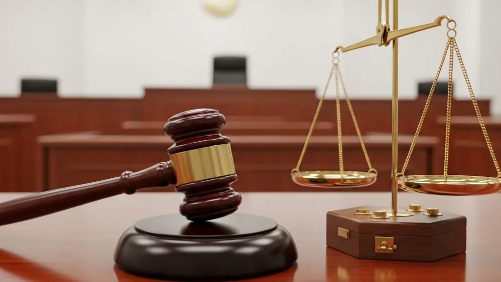 A large wooden gavel resting on a mahogany table next to a stack of balanced metal scales in a brightly lit modern courtroom setting.