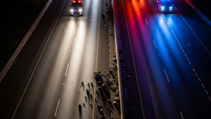 A high-angle view of a multi-lane highway at night with emergency vehicle lights reflecting off the asphalt and debris scattered near a concrete median.