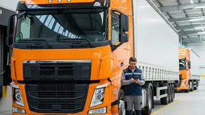 A large orange semi-truck parked in a logistics terminal with a driver standing nearby looking at a handheld device.