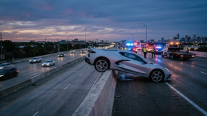 A silver Chevrolet Corvette sports car balanced precariously on the edge of a concrete freeway overpass with emergency vehicles and flashing lights in the background.