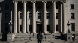 A person standing alone in front of a large government-style stone building with tall columns, looking up at the structure with a resolute expression; the lighting is dramatic with long shadows stretching across the plaza.