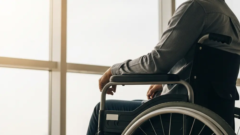 A close-up of a person in a wheelchair looking out of a large window towards a bright horizon, symbolizing the long-term journey of recovery and adaptation after a life-altering event.