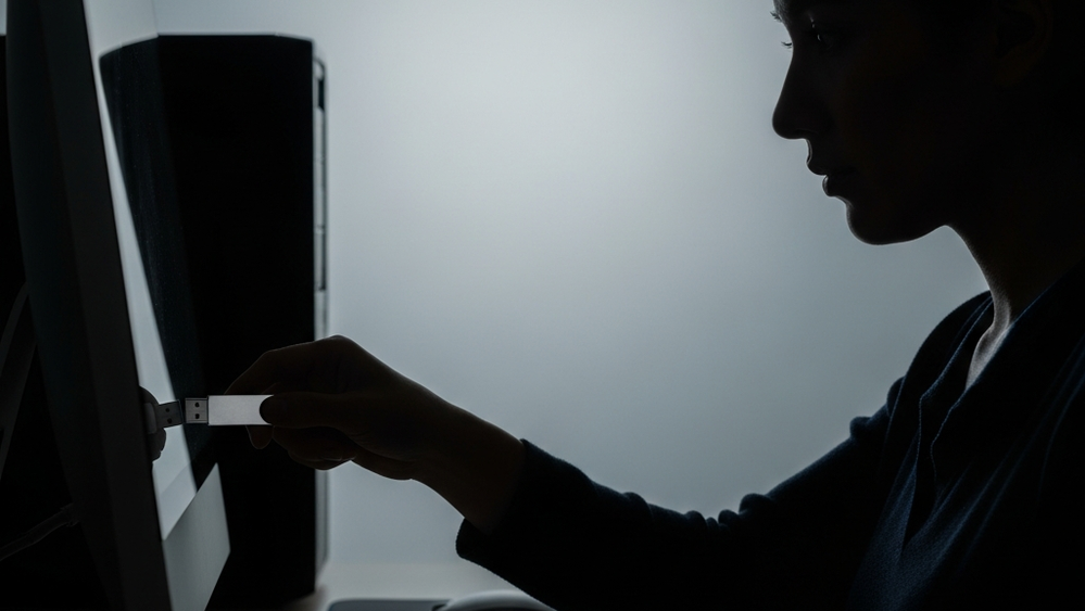 A silhouette of a person in a dark office using a USB drive on a computer workstation, symbolizing a digital data breach.