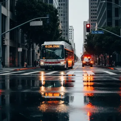 A modern city bus stopped at an urban intersection in downtown Los Angeles with emergency vehicle lights reflecting in the wet pavement