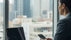 A person in professional business attire sitting at a modern desk with an open laptop and a smartphone, looking out a window at a city skyline.