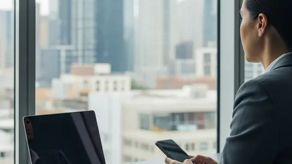 A person in professional business attire sitting at a modern desk with an open laptop and a smartphone, looking out a window at a city skyline.