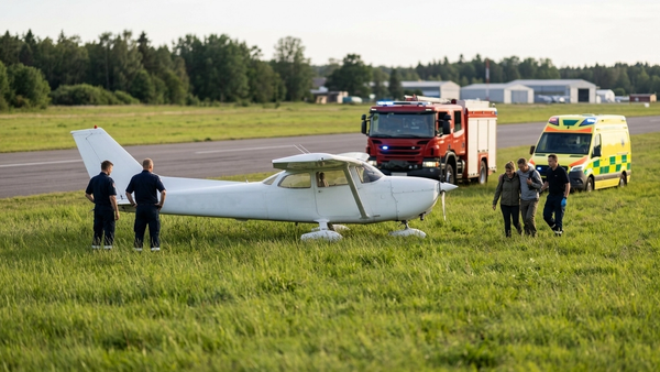 A small white propeller plane resting in a grassy field with emergency response vehicles in the background at an airfield.