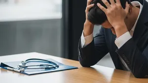 A person sitting at a wooden desk with their head in their hands looking stressed with a medical folder and a stethoscope nearby