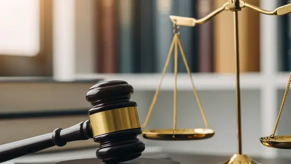 A close-up of a wooden gavel resting on a desk next to a balanced scale of justice, with blurred books in the background.