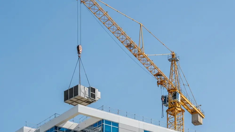 A yellow construction crane lifting a heavy HVAC unit onto a modern building rooftop under a clear blue Florida sky.