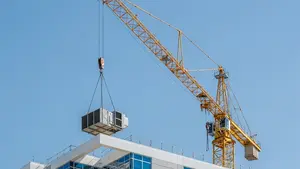 A yellow construction crane lifting a heavy HVAC unit onto a modern building rooftop under a clear blue Florida sky.
