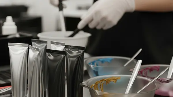 A close-up of several professional hair dye tubes and mixing bowls on a salon workstation, with a stylist in the background wearing protective gloves.