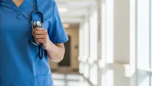 A healthcare professional in blue scrubs and a face mask holding a stethoscope while standing in a bright hospital hallway.
