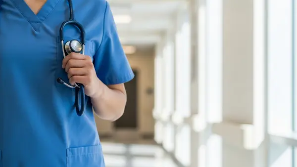 A healthcare professional in blue scrubs and a face mask holding a stethoscope while standing in a bright hospital hallway.