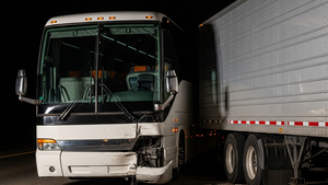 A large white tour bus with a damaged front end resting against the rear of a silver semi-truck trailer on a highway shoulder at night.