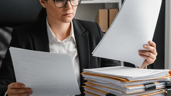 A person sitting at a desk looking frustrated while reviewing a stack of official documents and letters with a calculator nearby.