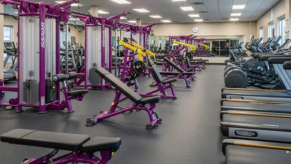 A large exercise facility with purple and yellow fitness machines, weight benches, and treadmills arranged in rows.