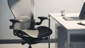 An empty ergonomic office chair sits at a modern white desk with a closed laptop and a ceramic mug in a sunlit, quiet office.