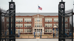 A large brick school building with a flag flying in front, seen through the iron gates of a playground with no people or text visible.