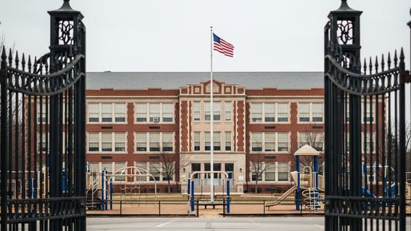 A large brick school building with a flag flying in front, seen through the iron gates of a playground with no people or text visible.
