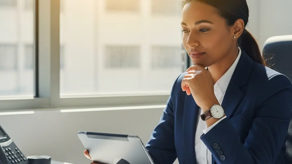A professional person in a business suit sitting at a modern desk, looking thoughtfully at a digital tablet while sunlight streams through a large window in a clean office setting.