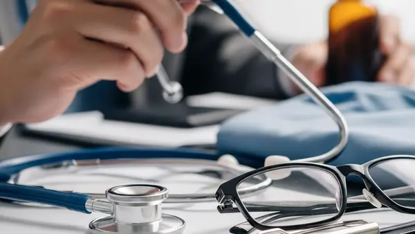 A close-up of a doctor's stethoscope resting on a desk next to a blurred-out medical folder and a pair of eyeglasses in a professional setting.