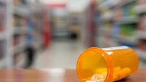 An orange prescription bottle with white pills spilling out onto a wooden pharmacy counter with a grocery store aisle blurred in the background.