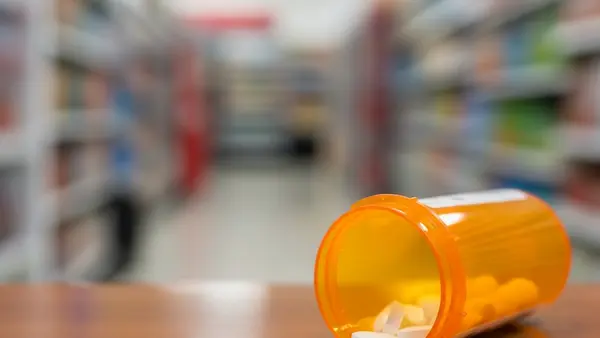 An orange prescription bottle with white pills spilling out onto a wooden pharmacy counter with a grocery store aisle blurred in the background.