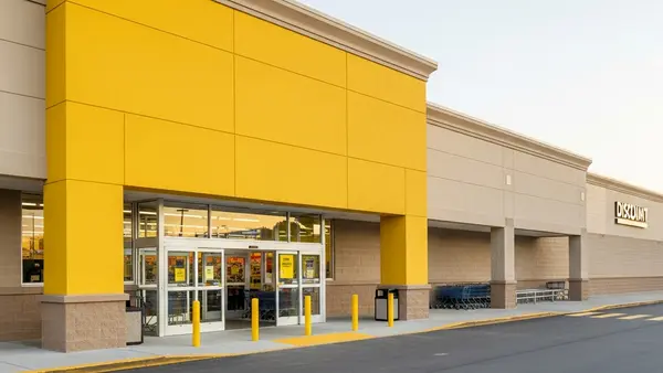 An exterior view of a discount retail store with a prominent yellow facade and automatic glass doors, situated next to a paved parking lot under a clear sky.
