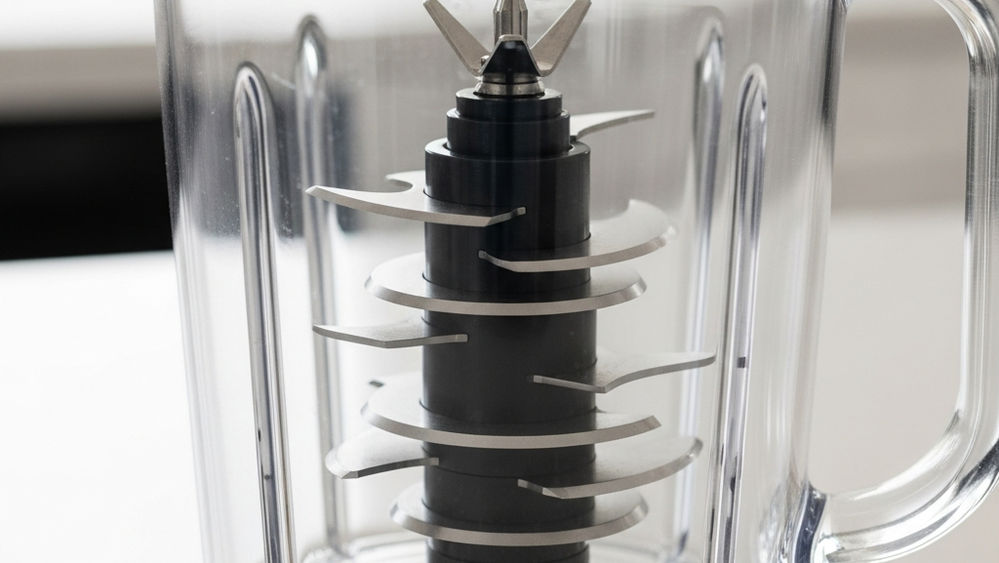 A high-speed kitchen blender sits on a counter, focusing on the sharp multi-tiered metal blade assembly inside a clear plastic pitcher.