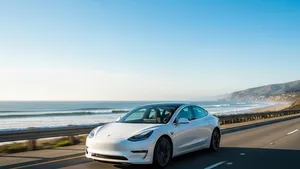 A white Tesla Model 3 sedan driving on a multi-lane coastal highway under a clear blue sky without any other cars nearby.