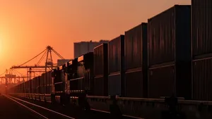 A long freight train moving along a industrial rail line during sunset with silhouettes of shipping containers