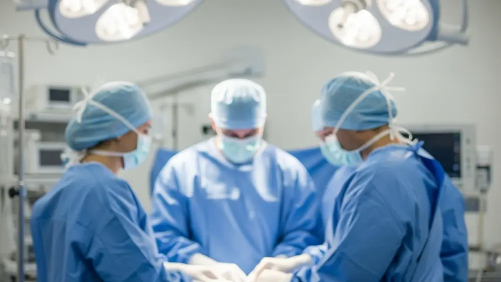 A blurred image of a medical surgical team in blue scrubs standing around an operating table under bright surgical lights, with medical equipment in the background.