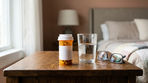 An amber prescription bottle labeled Gabapentin sitting on a wooden nightstand next to a glass of water and a pair of reading glasses.