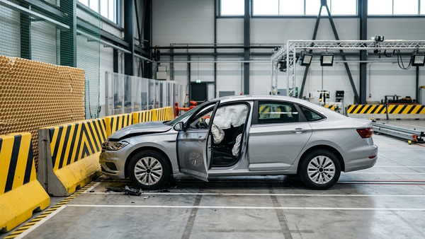 A modern silver sedan with a deployed driver-side airbag parked in a safety testing facility with crash test barriers nearby