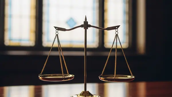 A symbolic pair of brass scales of justice standing on a mahogany desk in front of a soft-focus stained glass window background.