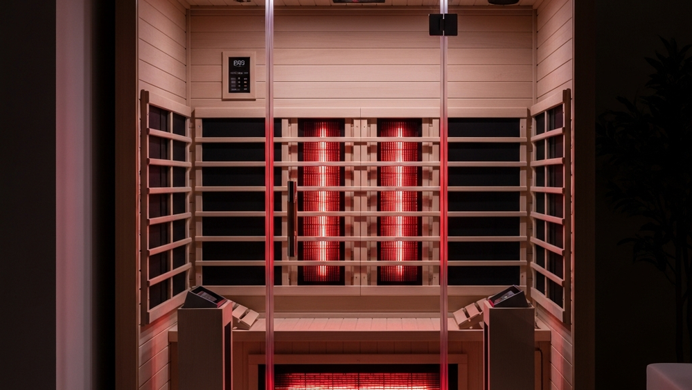 A modern infrared sauna with glass doors and wooden interior panels sits in a dimly lit room, with glowing red heating elements reflected in the glass door.
