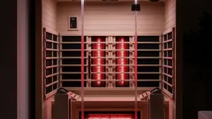 A modern infrared sauna with glass doors and wooden interior panels sits in a dimly lit room, with glowing red heating elements reflected in the glass door.